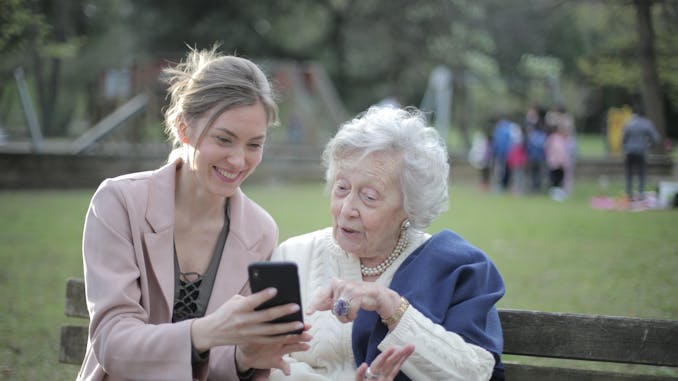 Delighted female relatives sitting together on wooden bench in park and browsing mobile phone while learning using