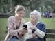 Delighted female relatives sitting together on wooden bench in park and browsing mobile phone while learning using